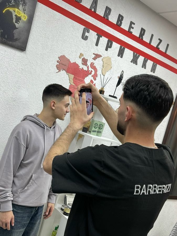 Барбер фотографирует готовую стрижку клиента у стены с логотипом BARBERIZI